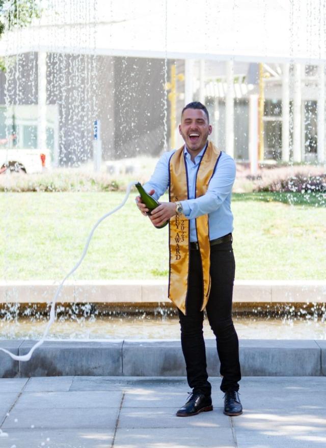 Gennaro Tecchia opening a bottle of champagne while wearing a graduation stole
